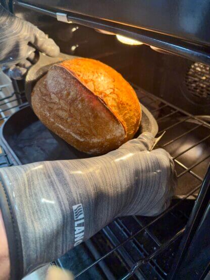 Oven gloves in use in an oven lifting a loaf of bread on a BreadMat out of a cast iron baker.